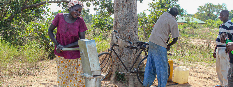 Nigerian woman pumps water from her community water pump while a man grabs something from his bike