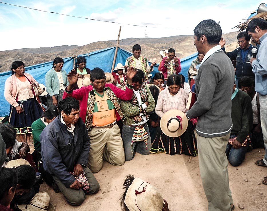 Bolivian men and women on their knees and others standing while two missionaries stand in front of them and pray
