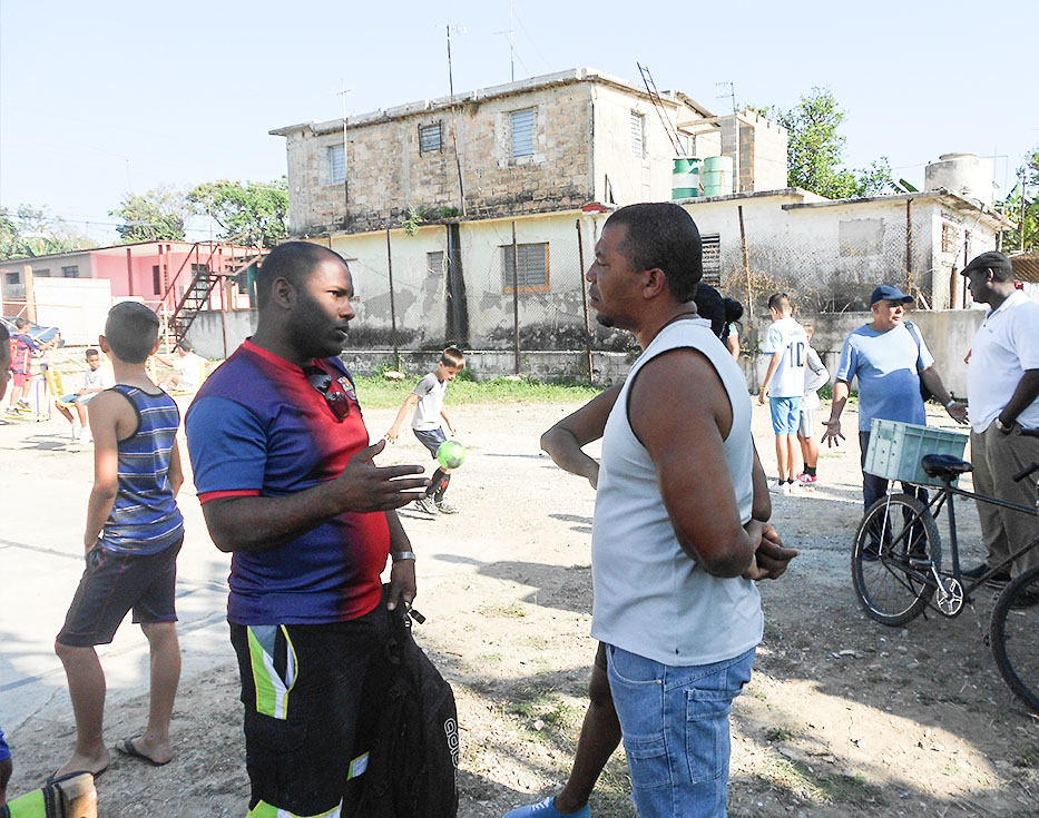 Two Cuban men talking while children play soccer and other adults stand around