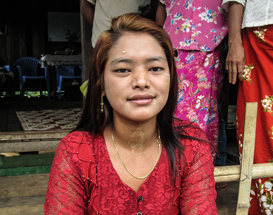 Burmese girl wearing a red dress sitting on the steps of her house outside with other women behind her