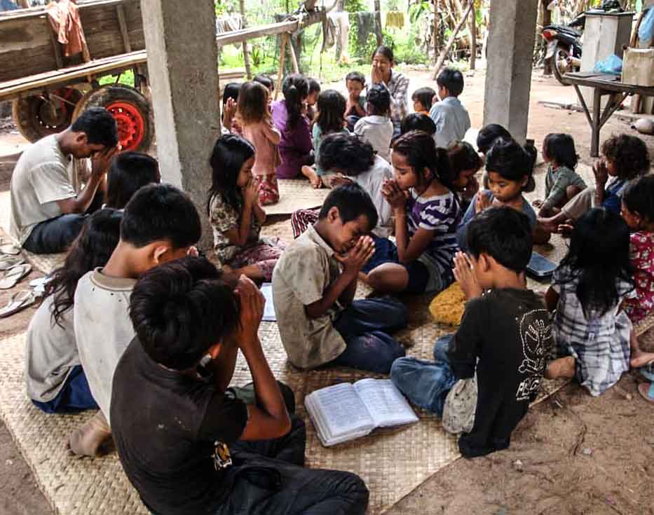 School children sitting under a concrete pavilion with their hands together praying