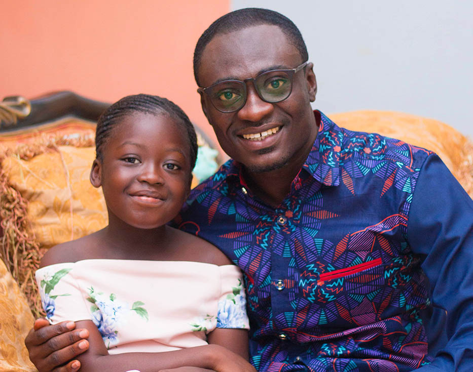 Ghanaian father sits on couch smiling with his arm around his daughter