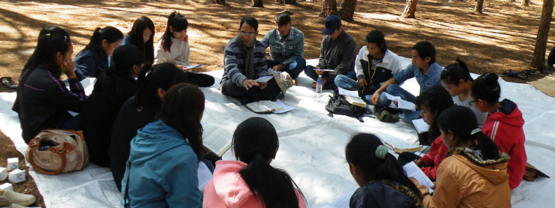 Christian missionary leads Bible study of young adult Christians who sit in a circle on the ground