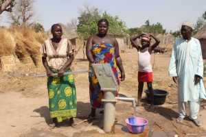 Nigerian woman pumps water from community well into large bowl while another woman, boy, and man watch