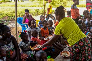 Sierra Leonean woman hands two bowls of food to children sitting with their mother while other children are eating