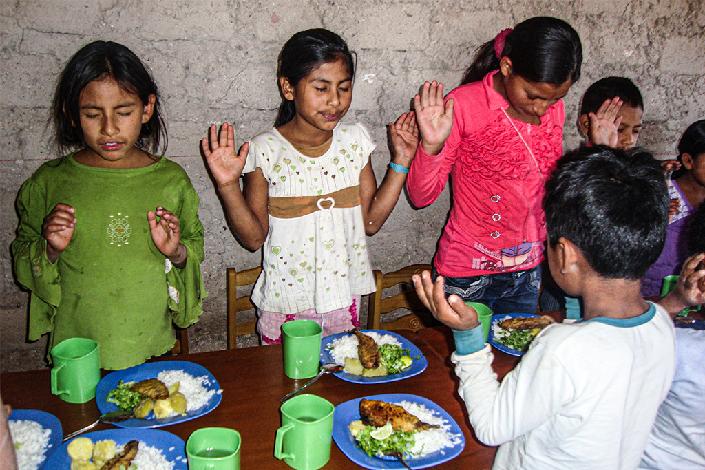 Latin American children stand praying before they eat their dinner on the table in front of them
