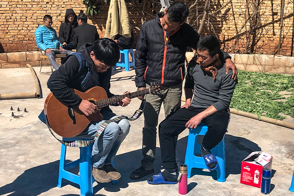 A Chinese man sits on a stool playing guitar while two other Chinese men sit and stand nearby listening and watching