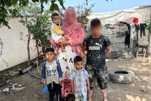 Middle Eastern mother stands in a refugee camp with her three boys while holding her youngest son