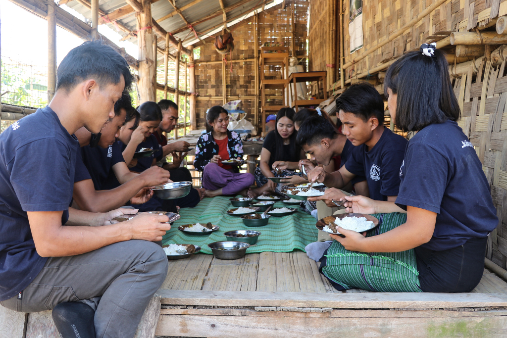 Burmese young adults sitting cross-legged on a wooden floor under a pavilion made from bamboo eating rice for lunch