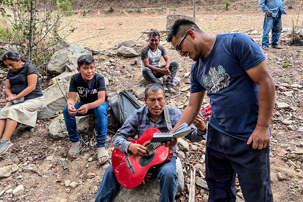 Mexican man holds songbook for another man playing a red guitar while others sit on the ground around them and listen