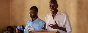 Sudanese Christian pastor wearing a white dress shirt preaches to his congregation from the pulpit with another brother standing next to him