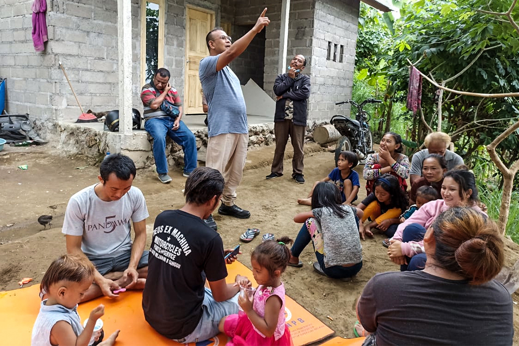 Indonesian missionary shares a Bible story with children and parents as they sit outside on a dirty path