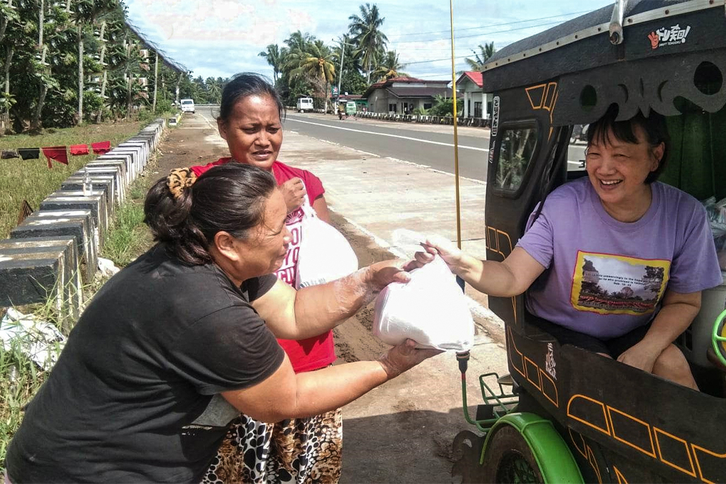 Filipino woman in a motorized cart hands a bag of food to a woman on the side of the road while another woman holds a different bag of food