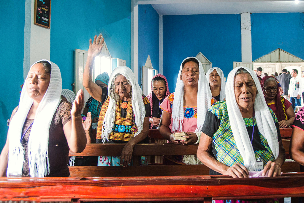Mexican Christian women stand in front of wooden pews with their eyes closed and hands raised praising God