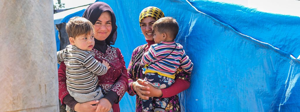 Two Afghani refugee women standing in a refugee camp each holding their young son and smiling
