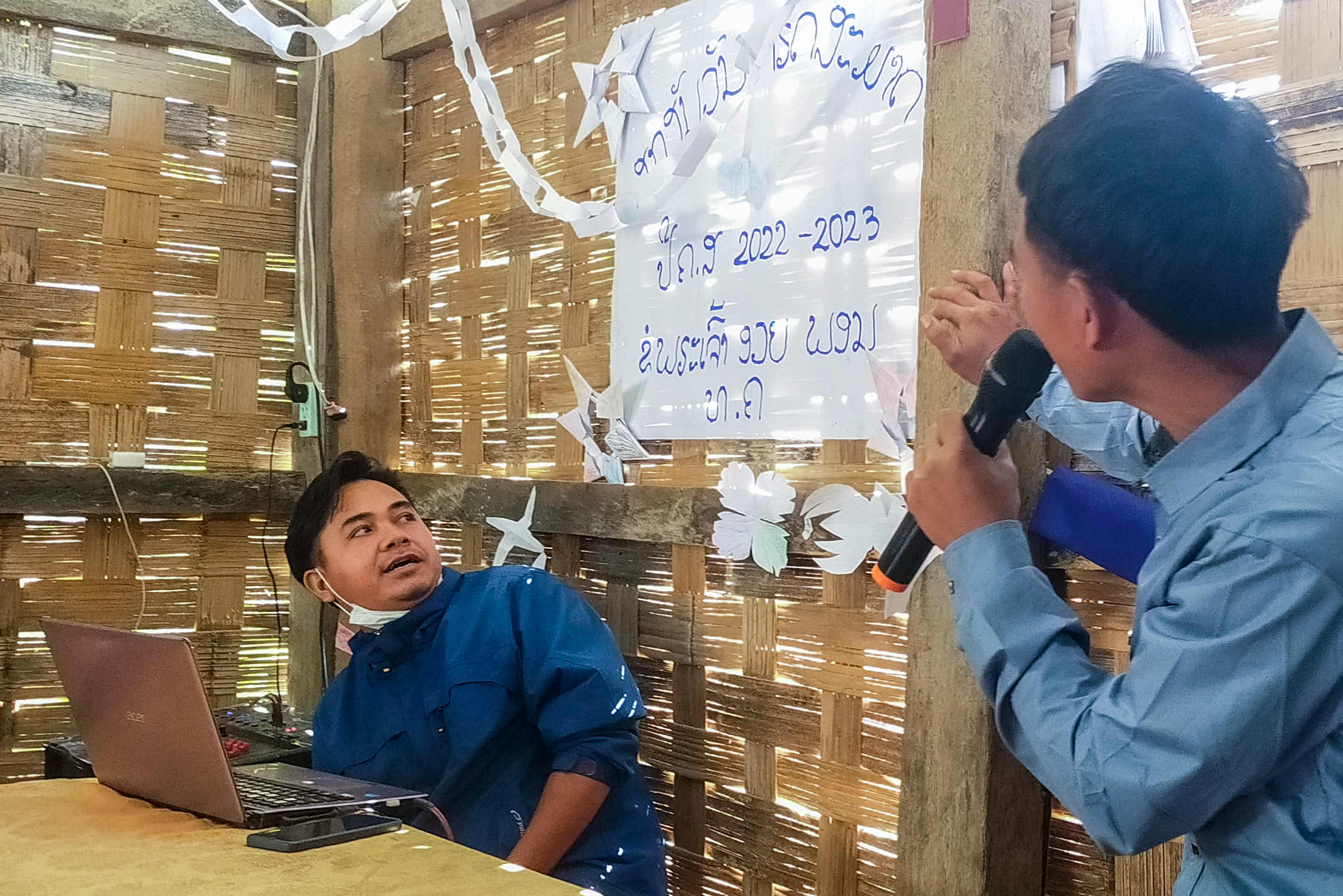 Laotian Christian points a sign while speaking through a microphone while another man works on the electronic equipment