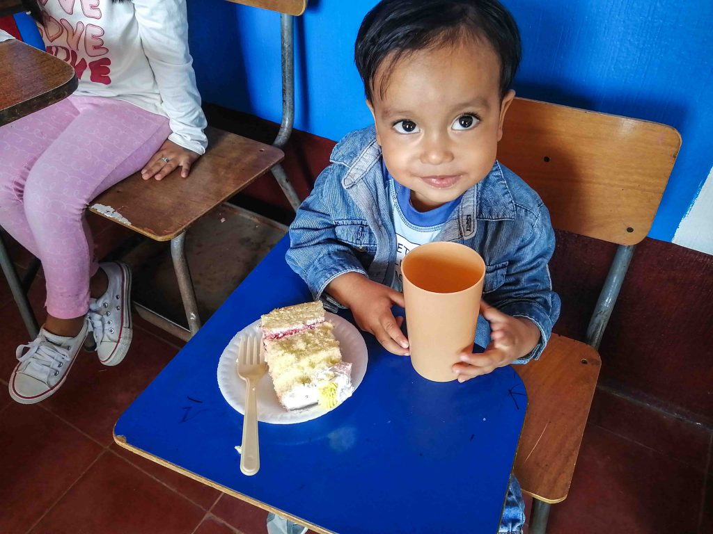 A Guatemalan child sitting in at a school desk eating a piece of cake holding a plastic cup