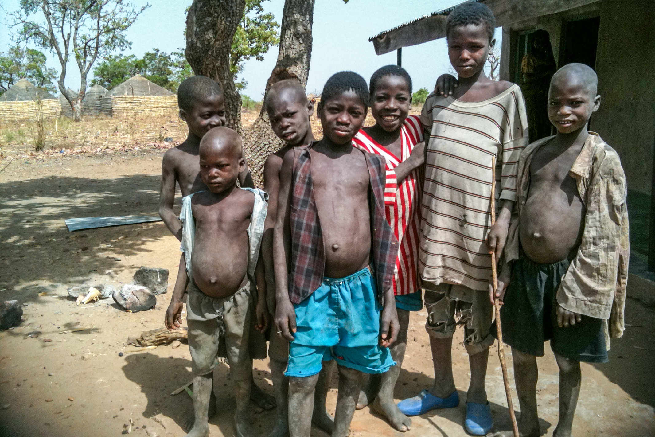 7 Nigerian boys standing outside of their house wearing tattered clothes and their legs covered in dirt
