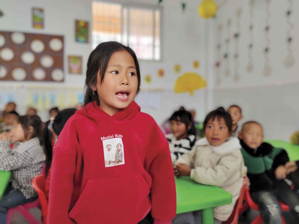 Chinese little girl standing in a classroom with the rest of her classmates sitting behind her at green tables