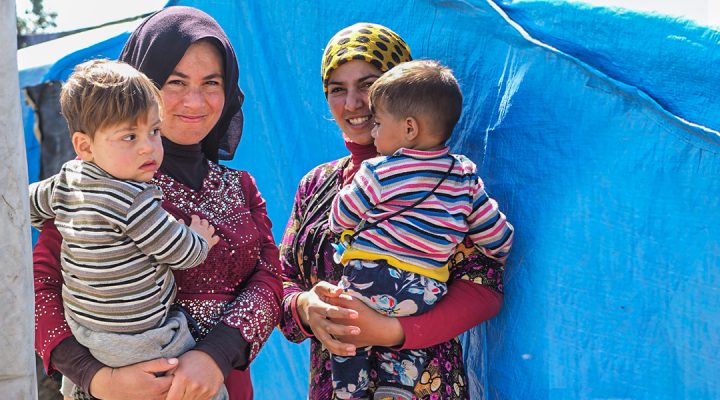 Two Afghani refugee women standing in a refugee camp each holding their young son and smiling